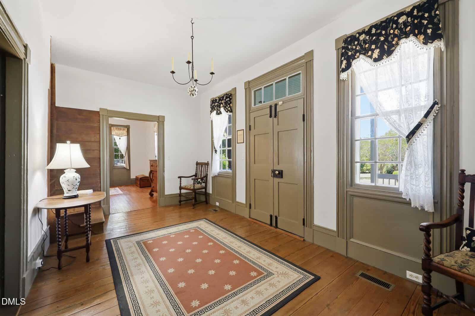 Bright hallway with wooden floors, beige trim, a patterned rug, antique chairs, a small table with a lamp, and large windows with lace curtains letting in natural light.