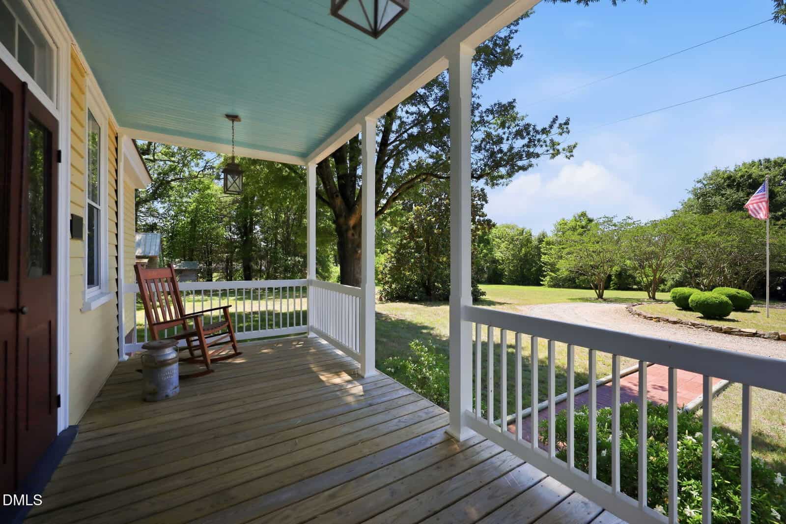 A wooden front porch with white railings, a light blue ceiling, two rocking chairs, and a milk can. The porch overlooks a yard with green trees, bushes, and an American flag on a sunny day.