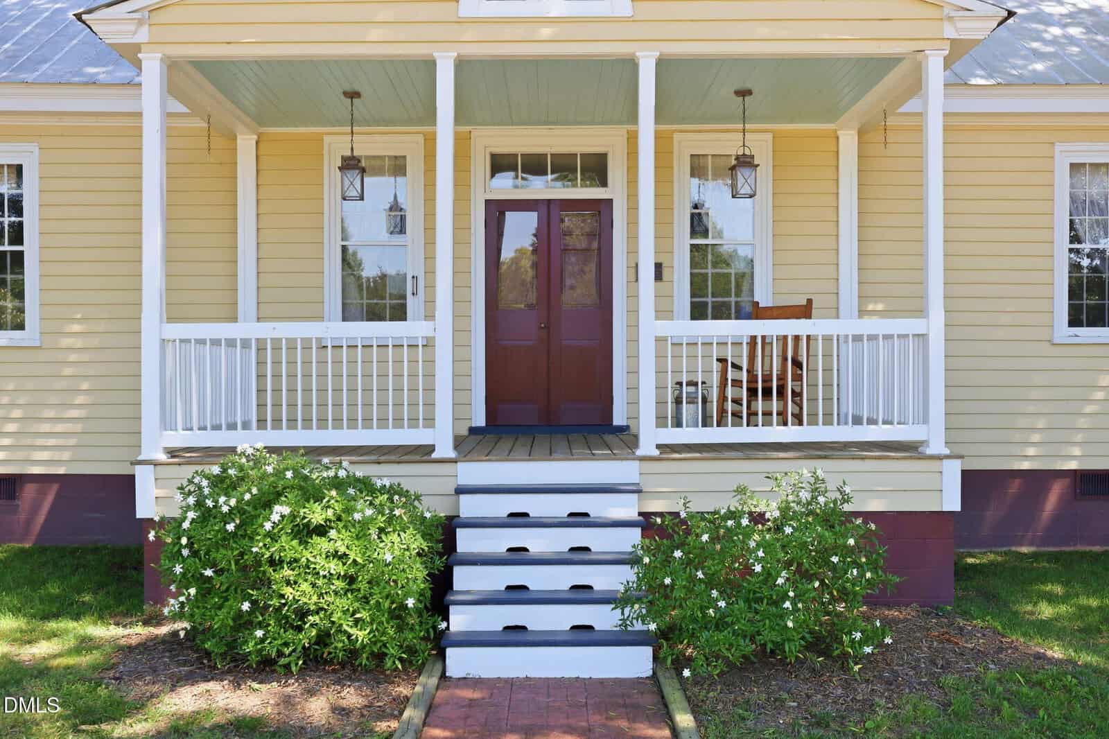 A yellow house with a covered front porch, white railings, and steps leading up. There are two hanging lanterns, a wooden rocking chair, and bushes with white flowers on either side of the steps.