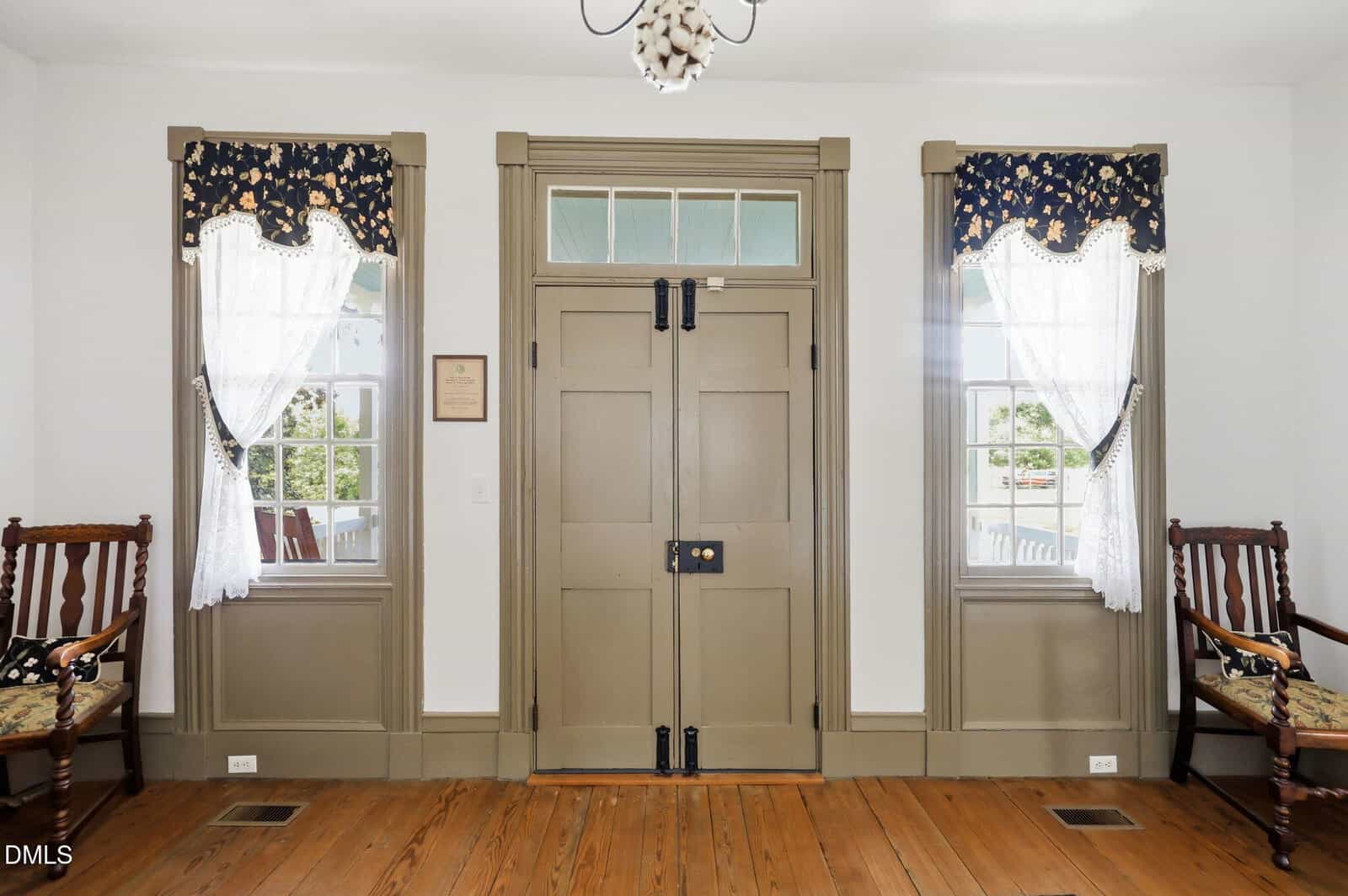 Front entryway with a taupe double door, two windows with lace curtains and floral valances, wooden trim, and two wooden chairs with patterned cushions on either side, all on a polished wood floor.