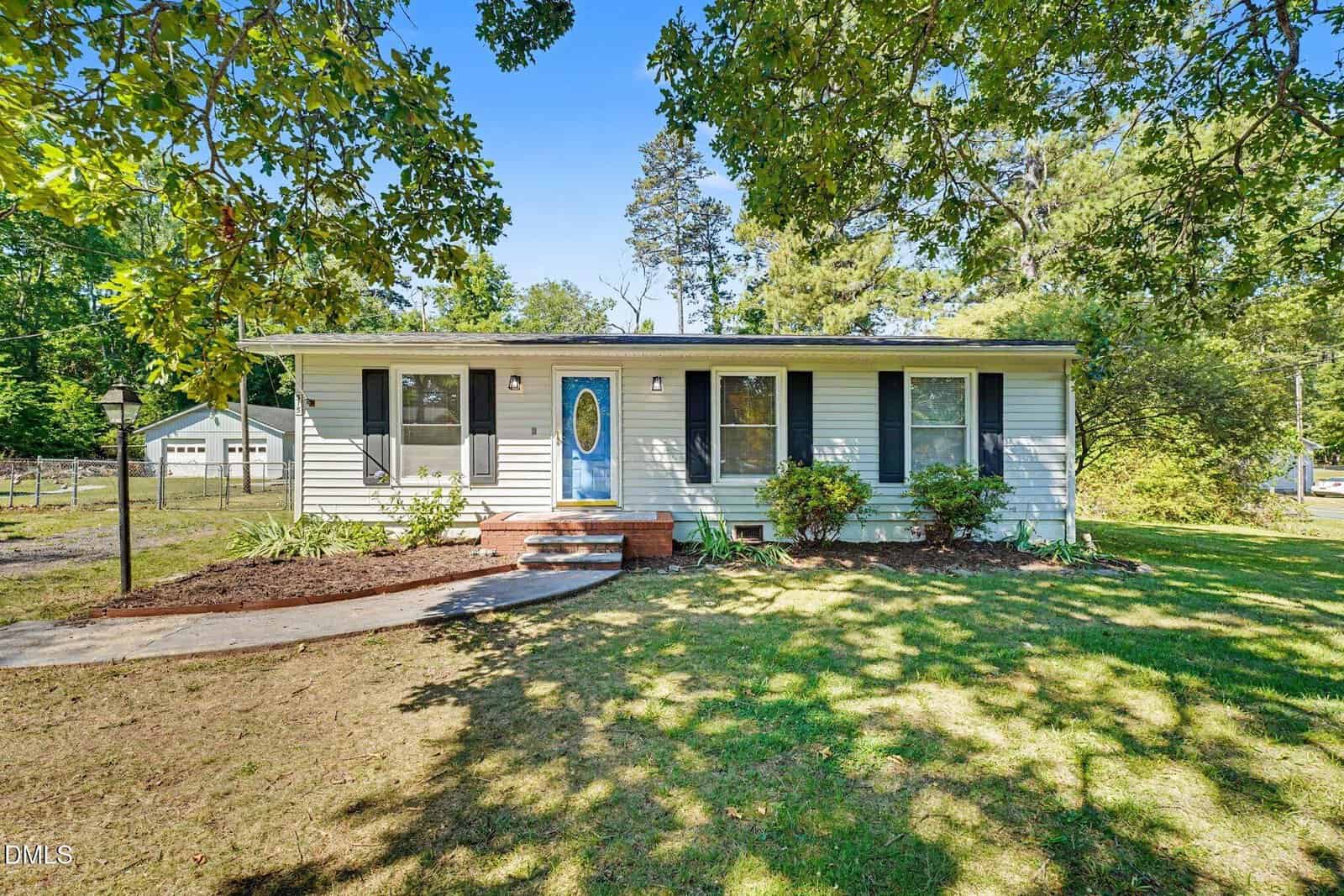 A single-story house at 315 6th Street in Butner features light-colored siding, a blue oval door, black shutters, and several front windows. The yard has green grass, shrubs, a curved walkway, and is shaded by large leafy trees.
