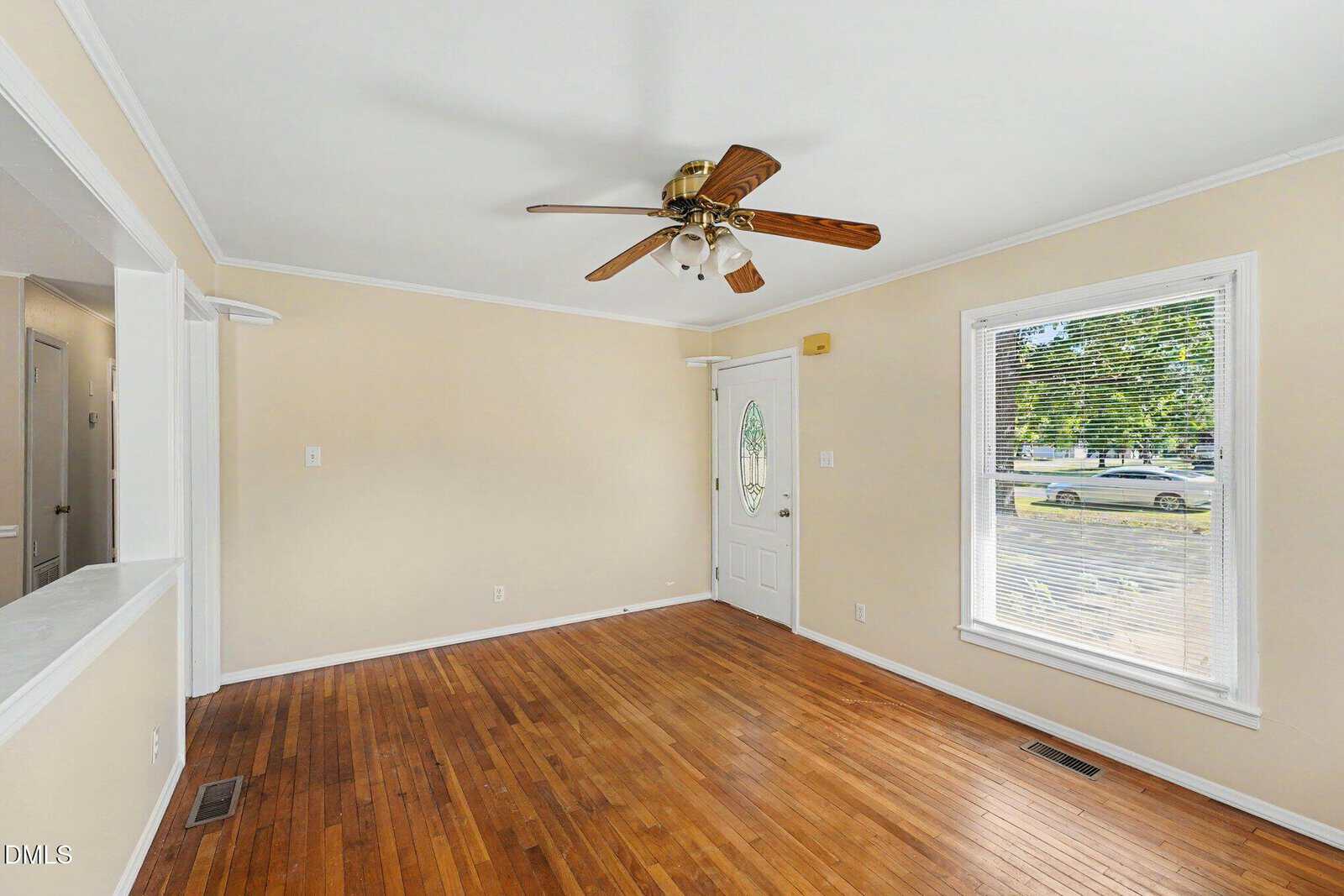 A bright, empty living room with light beige walls, wooden floors, a ceiling fan with lights, a white front door with an oval window, and a large window letting in sunlight.