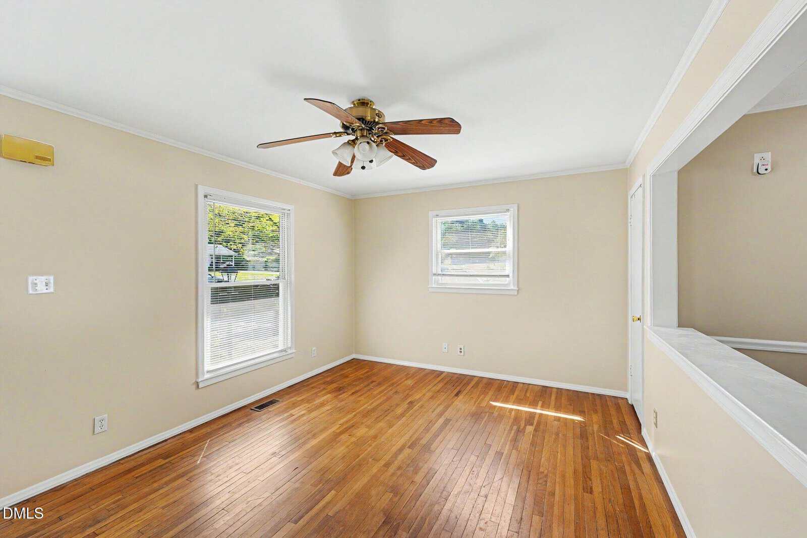 A bright, empty room with hardwood floors, beige walls, white trim, two windows, a ceiling fan with wooden blades, and an open pass-through to an adjacent space. Sunlight streams in from the windows.