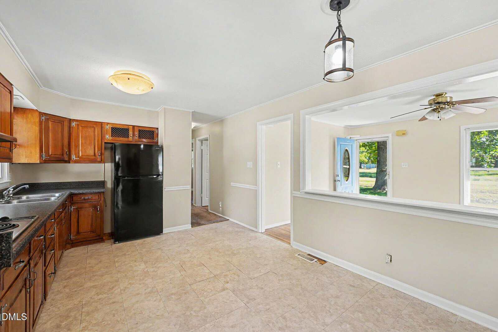 Bright kitchen with wooden cabinets, black appliances, and beige tile floor. Open window space looks into a living area with ceiling fan, large windows, and a view of the front door and green yard outside.