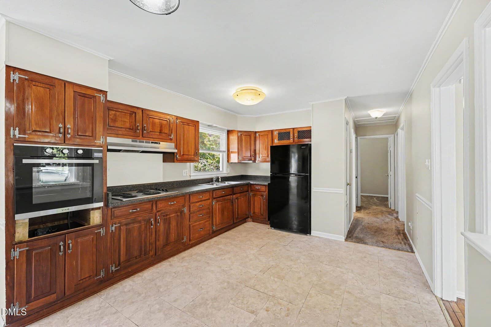 A kitchen with brown wooden cabinets, black appliances including a refrigerator and oven, granite countertops, tiled floor, and neutral-colored walls. A hallway is visible to the right.
