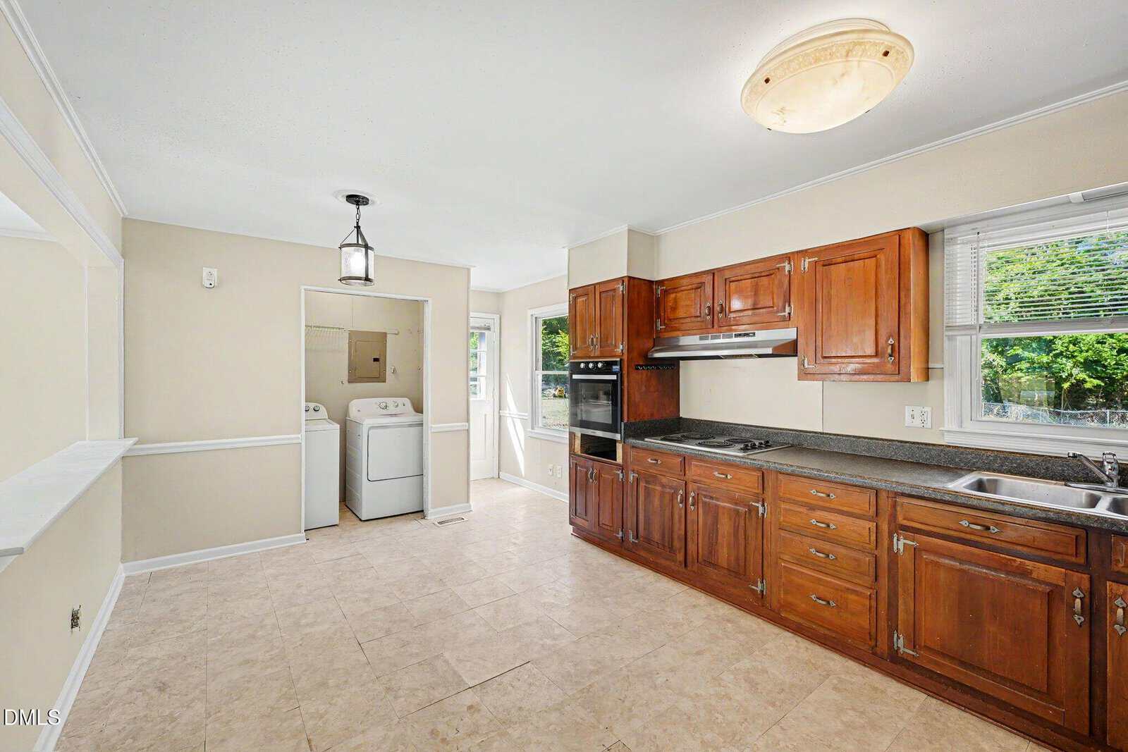 Bright kitchen with brown wood cabinets, black countertops, stainless oven, and double sink. Adjacent laundry area with washer and dryer. Large window lets in natural light. Cream-colored tile floor.