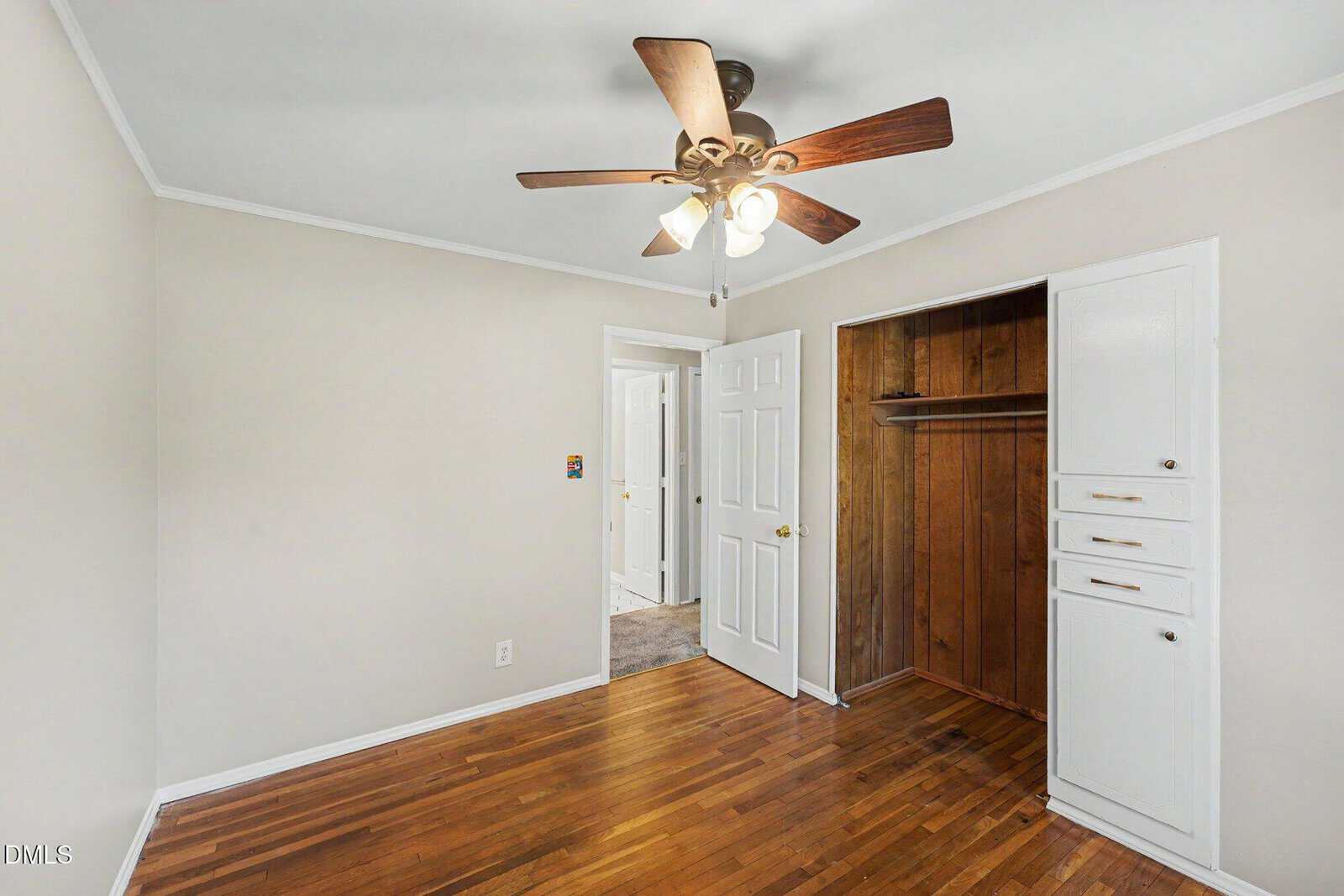 Small room with hardwood floors, beige walls, a ceiling fan with lights, white closet doors, built-in drawers, and an open section revealing wood paneling inside the closet. Doorway leads to a bright hallway.