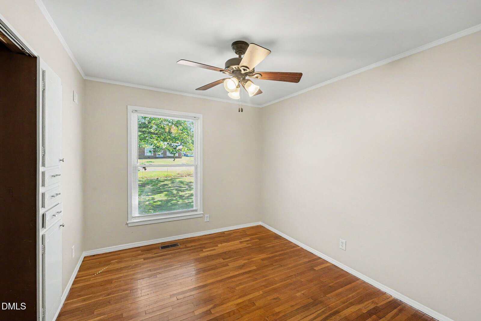 A small empty room with hardwood floors, beige walls, white trim, a window with blinds showing green trees outside, a ceiling fan with lights, and built-in cabinets on the left.
