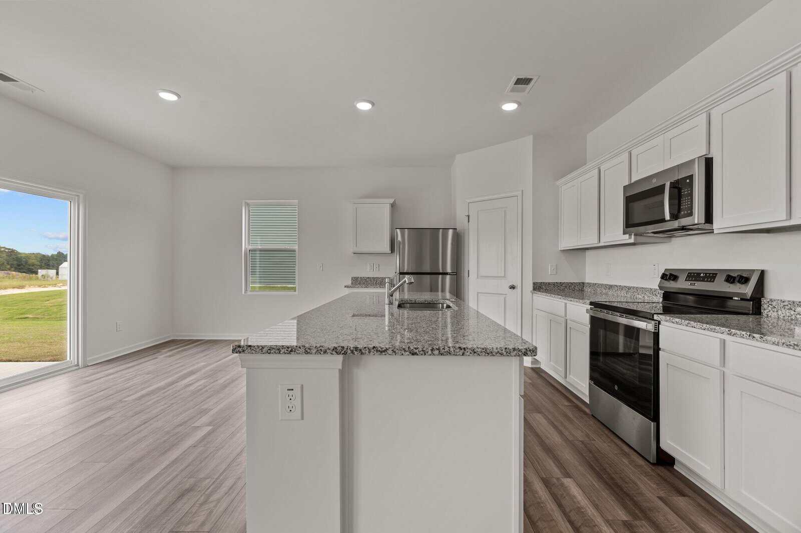 Modern kitchen with white cabinets, stainless steel appliances, granite countertops, and an island. The room has wood-style flooring, large windows, and a door leading to an outdoor area with a grassy view.