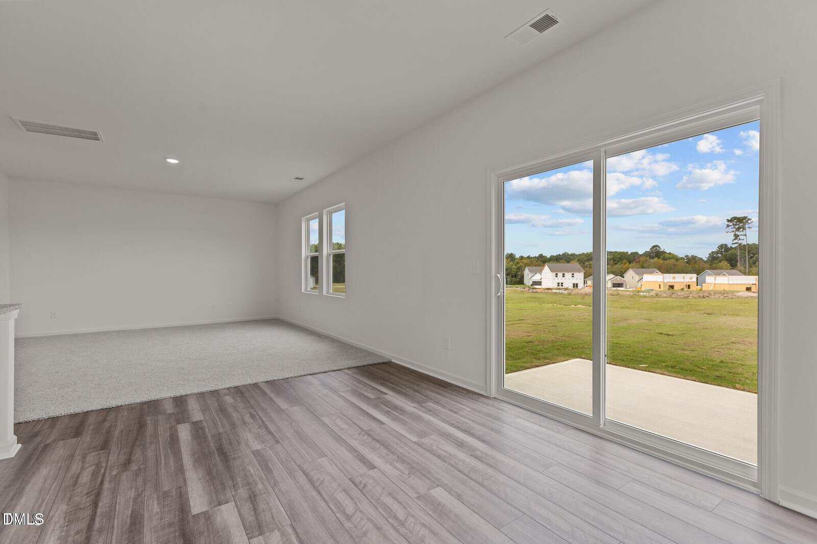 A bright, empty room with light gray wood flooring and carpeted area, large windows, and a sliding glass door opening to a grassy backyard with houses and trees in the distance under a blue sky.