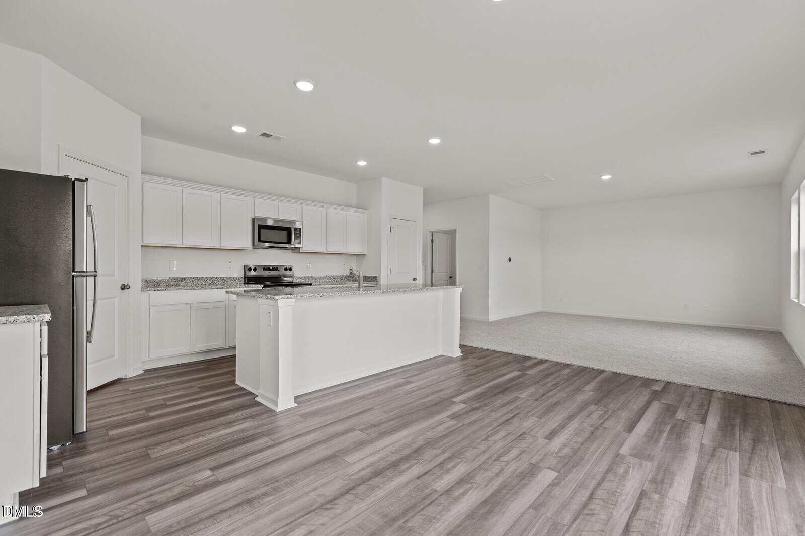 A modern open-concept kitchen with white cabinets, granite countertops, stainless steel appliances, and an island, opening to a spacious living area with carpeted flooring and large windows.