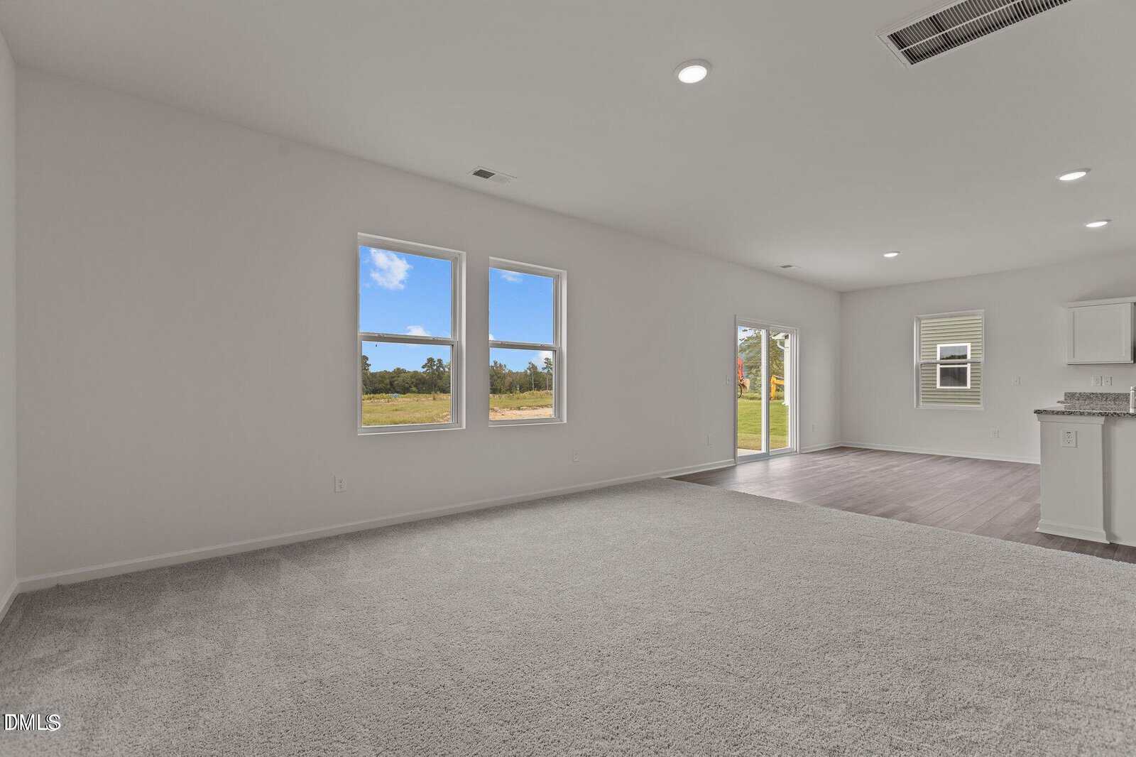 Bright, empty living room with carpet, large windows, and an open floor plan connected to a kitchen with white cabinets and wood flooring. Sliding glass doors lead to a grassy outdoor area.