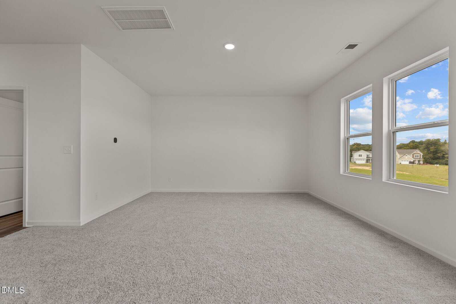 Empty room with white walls, light gray carpet, two large windows showing a green landscape, a closed white door, and a ceiling light. The room is well-lit with natural sunlight.