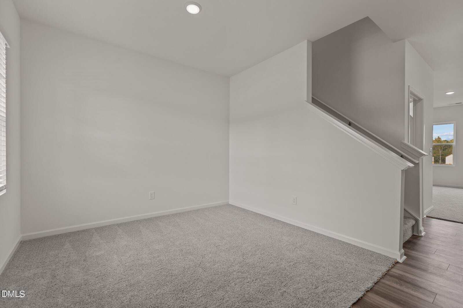 A bright, empty room with light gray carpet, white walls, and a small staircase leading up. Sunlight enters through a window on the left. The adjoining hallway has wood flooring.