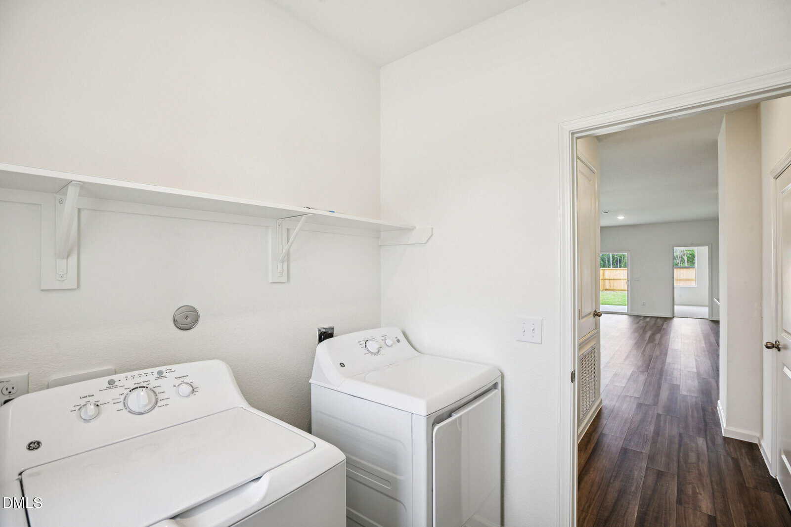 A laundry room with a white washing machine and dryer, a white wall shelf above them, and a door leading to a room with wood flooring and a view of a fenced backyard through large windows.