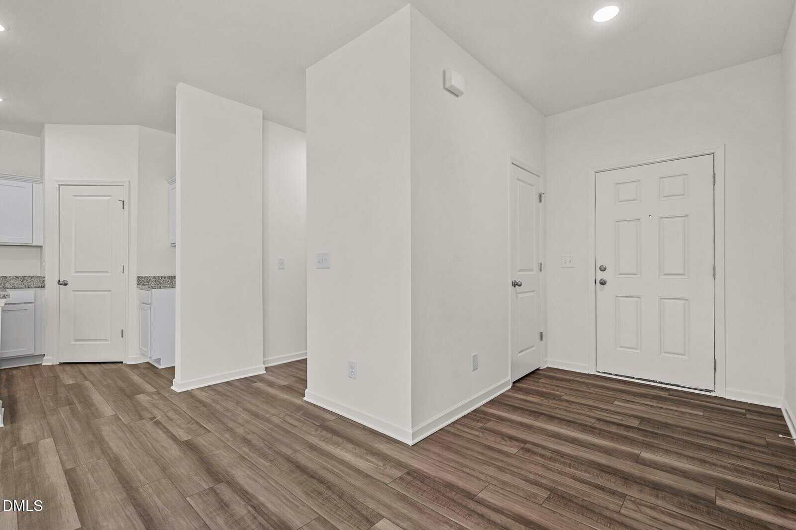 Bright entryway with white walls, recessed lighting, and wood-style flooring. A white door is visible on the right, with a view into an adjacent kitchen area on the left. Clean and modern interior design.