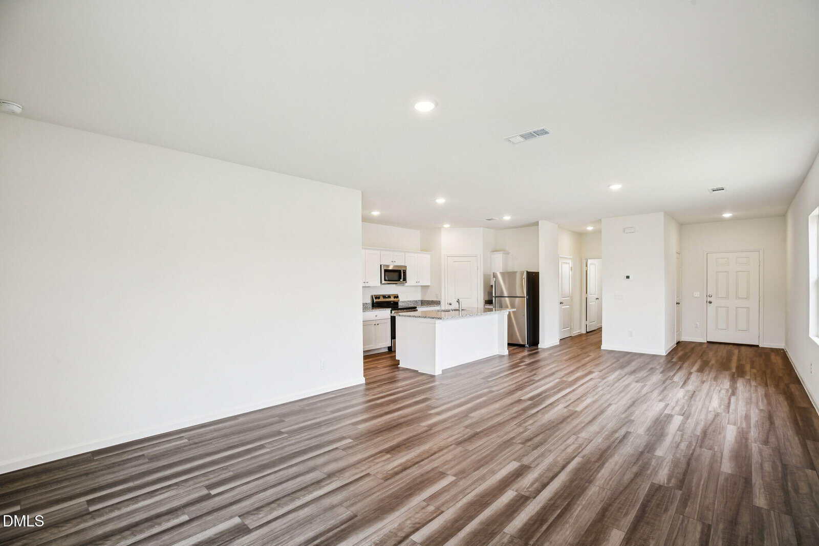 Open-plan interior with wood-style flooring, white walls, and recessed lighting. Visible are a kitchen with white cabinets and stainless steel appliances, and a living area with large windows and a door.