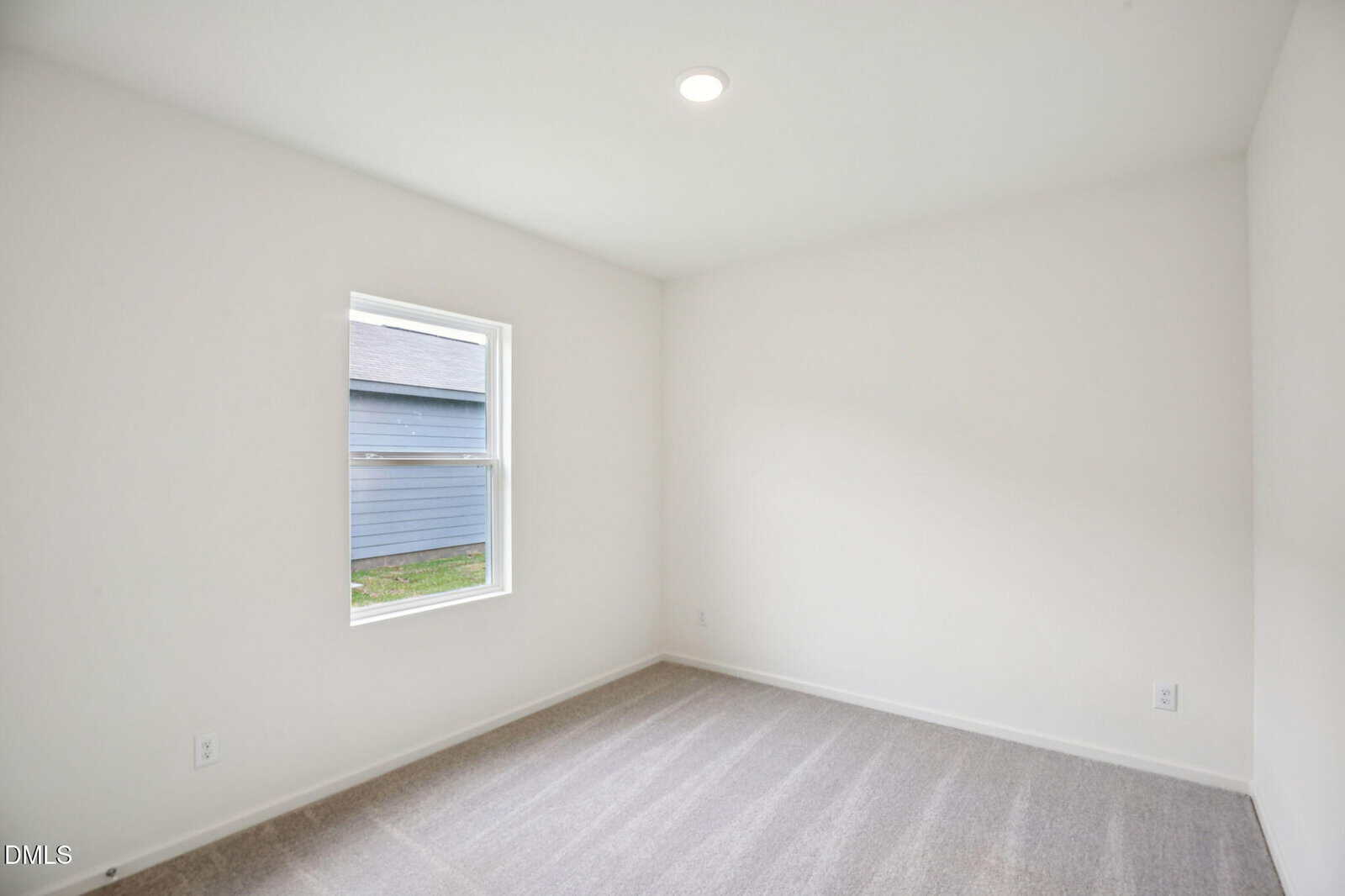 An empty room with white walls, beige carpet, a single window, and a ceiling light. The window shows the side of a neighboring building with blue siding. The room appears clean and well-lit.