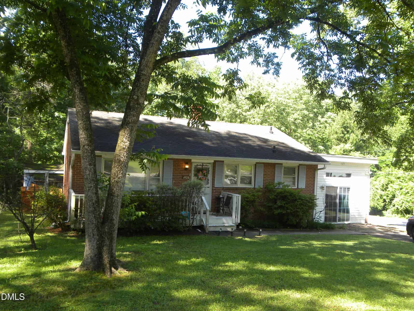 A one-story brick house with a porch and large windows sits at 1025 Carroll Road in Henderson, surrounded by green trees and bushes. The grassy lawn is shaded as sunlight filters through the leaves, and a driveway appears on the right side.