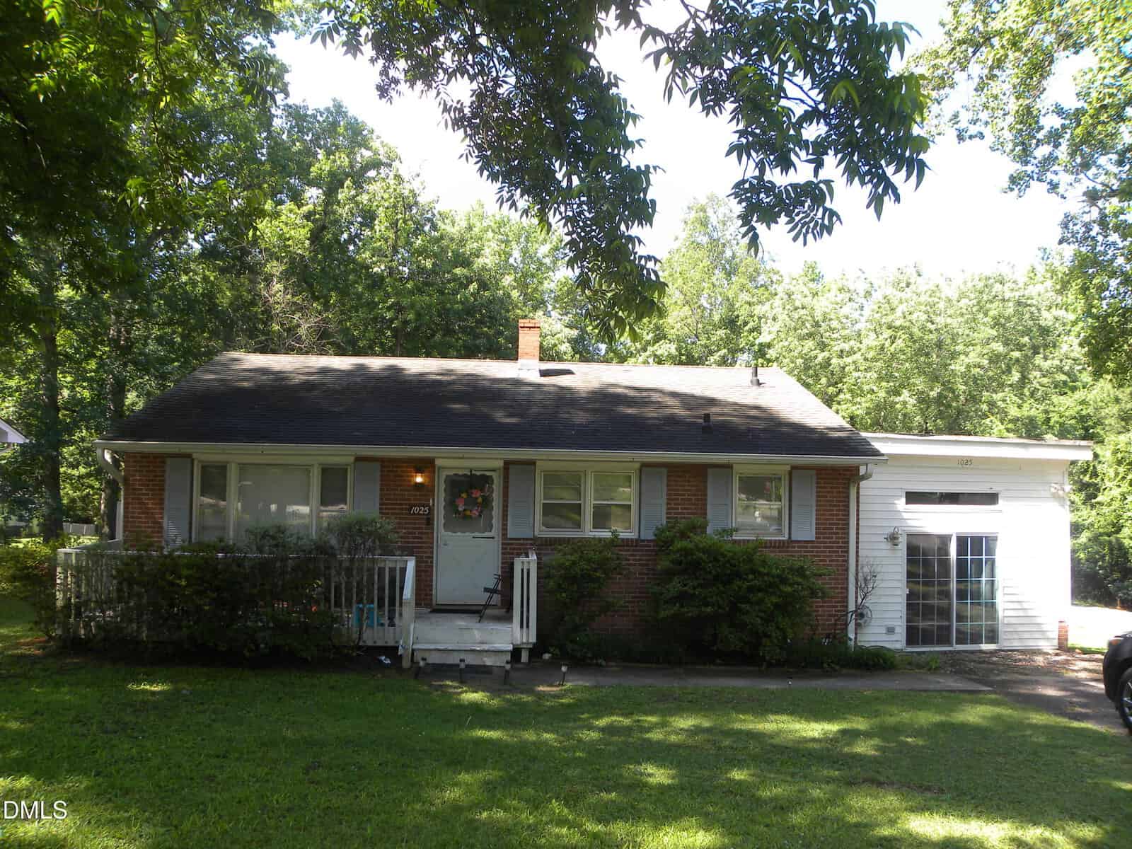 A small, single-story brick house with a front porch, white trim, and an attached white addition. The home is surrounded by green grass, bushes, and large leafy trees under a sunny sky.