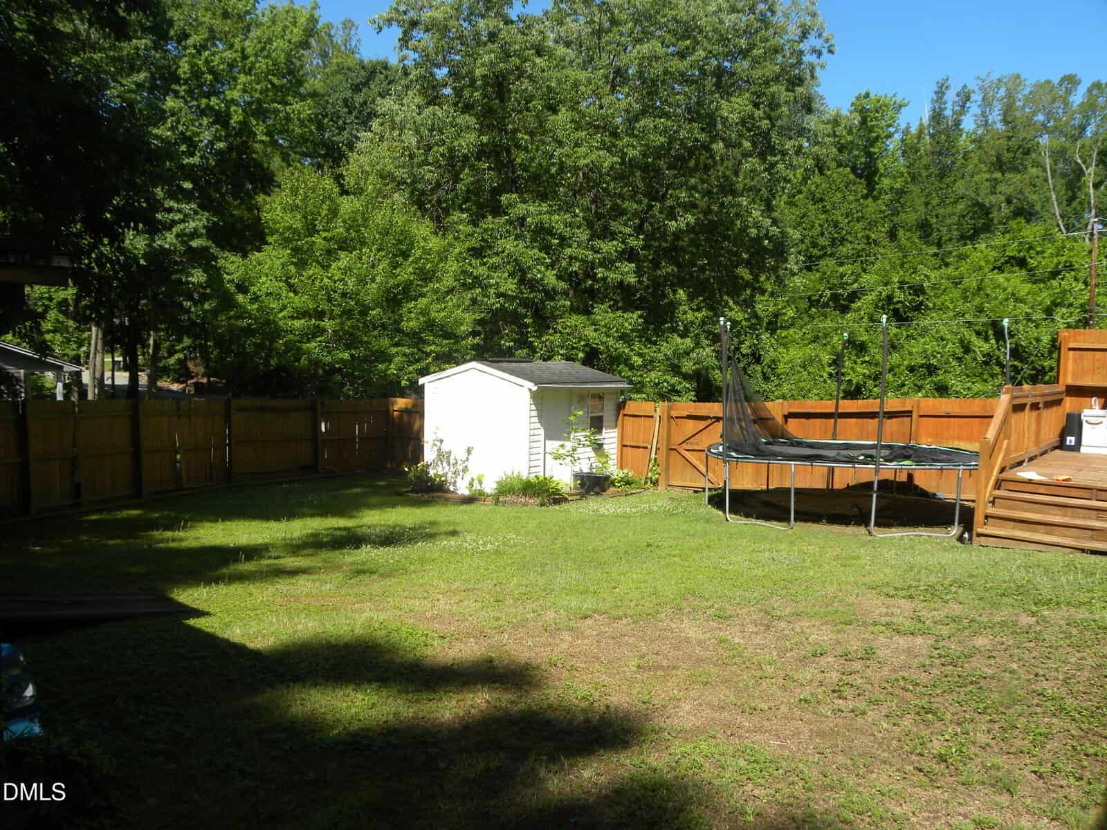 A fenced backyard with a grassy lawn, a small white shed, and a trampoline. There are trees in the background and sunlight casting shadows on the grass.