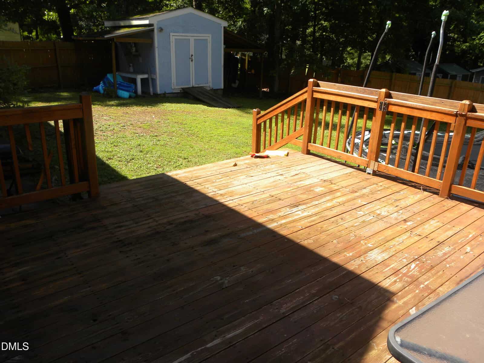 A sunlit wooden deck with stairs leads to a grassy backyard. In the background, there is a light blue shed and a wooden fence enclosing the yard. Shadows from the deck railing fall across the boards.