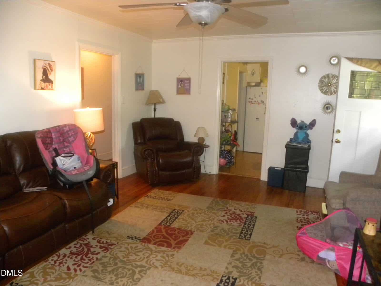 Living room with brown leather furniture, patterned rug, wall art, and a ceiling fan. A pink car seat is on the couch, toys are scattered, and a large Stitch plush sits near the doorway to the kitchen.