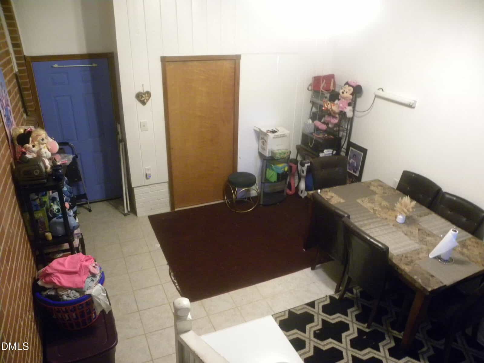 A dining room with a table and six chairs, kids’ toys on shelves, a blue door, a brown rug, and a laundry basket. The space is tidy and well-lit, with white walls and various storage items.