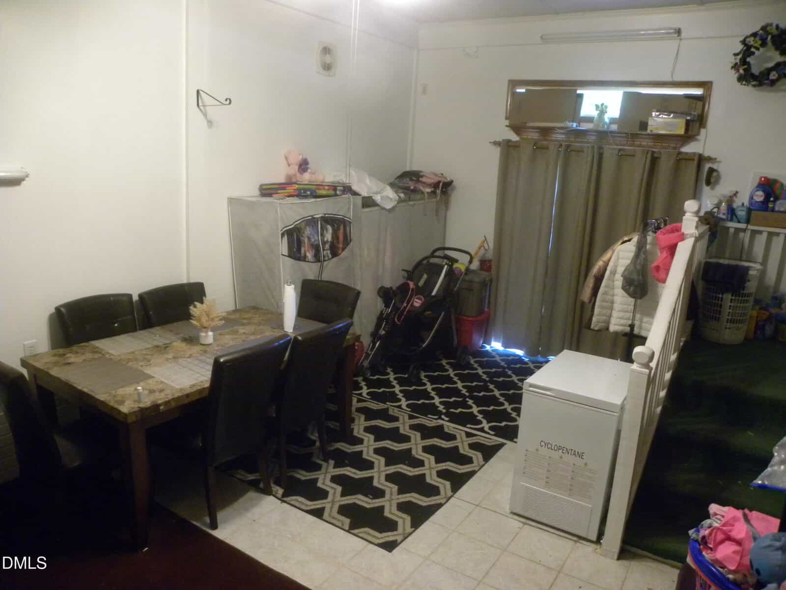 Dining area with a dark table and six chairs, storage bins, golf clubs, a rug, a washing machine, and curtains covering a sliding glass door. Jackets and laundry items are visible throughout the room.