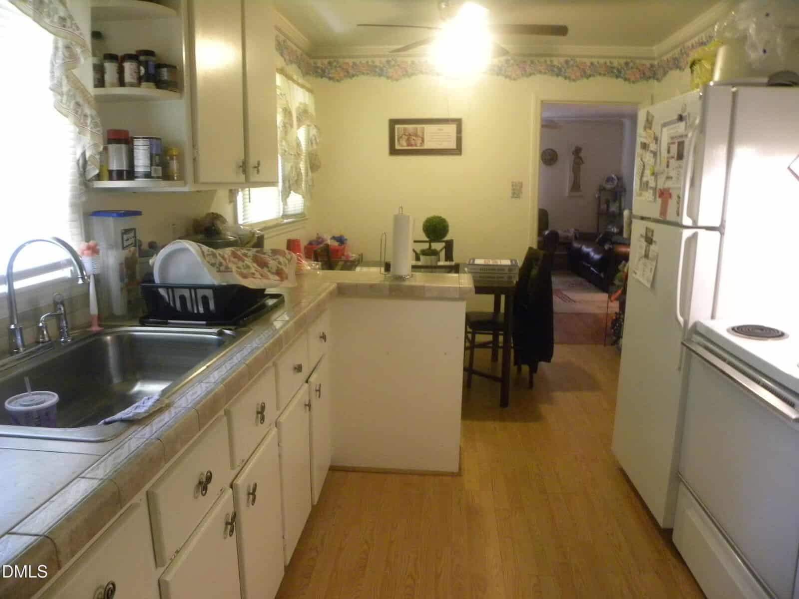 A narrow kitchen with white cabinets, a double sink, dish rack, and a refrigerator. There are shelves with spices and a small dining table is visible toward the back near an adjoining room.