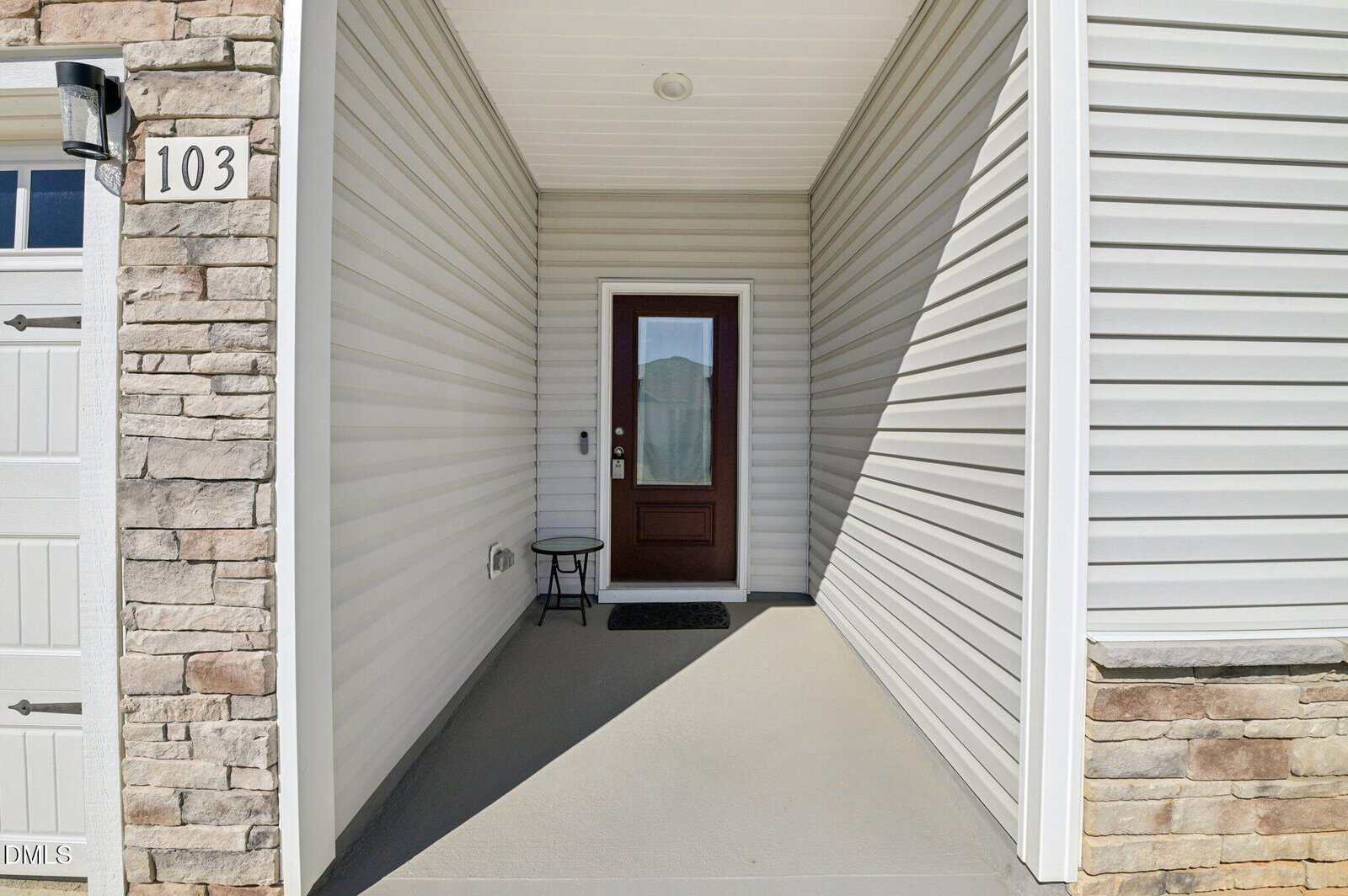 Covered front porch with light gray siding, a single maroon and glass front door, a small black table, stone accents, and the house number 103 next to the garage.