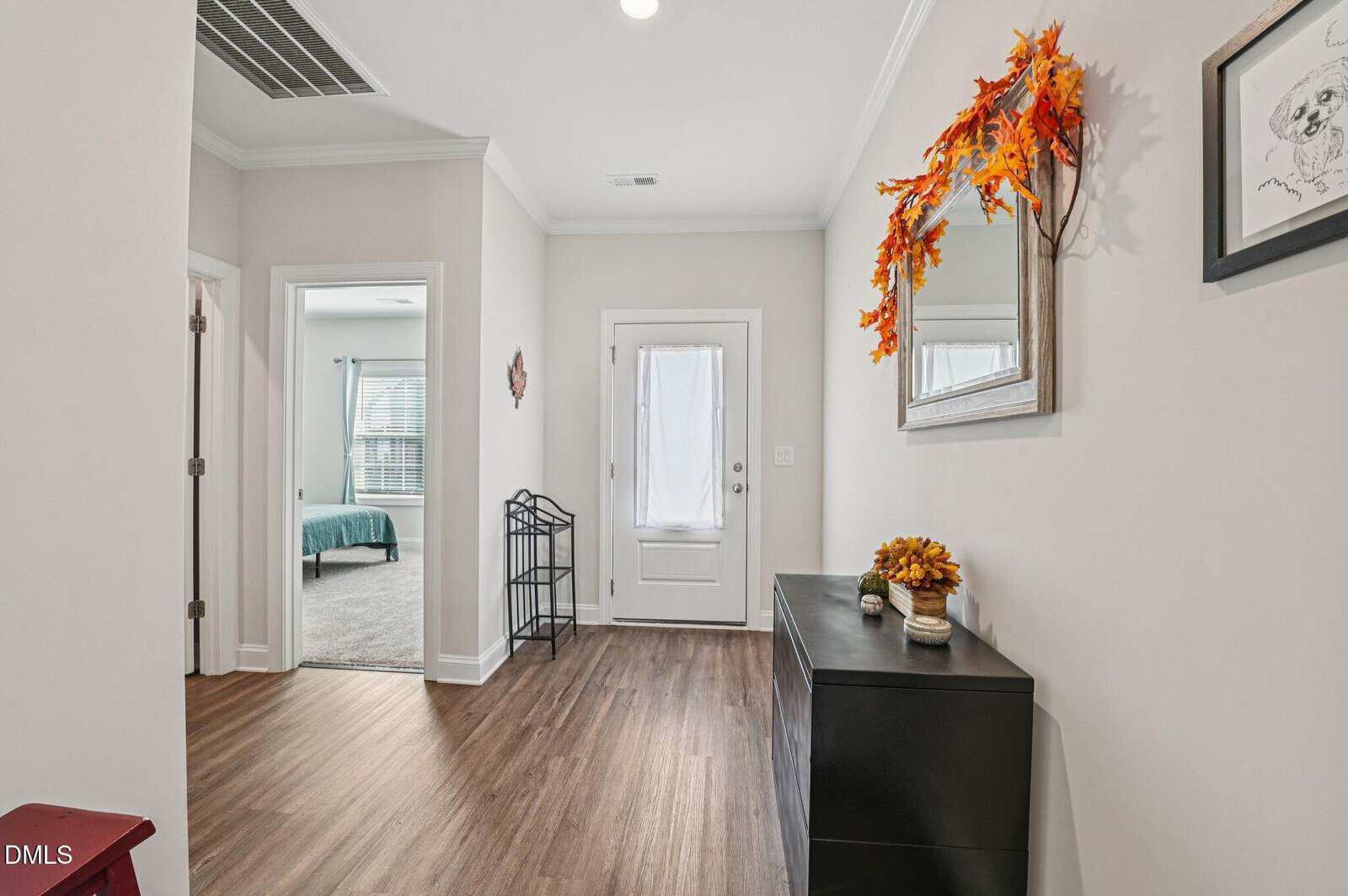 A bright hallway with wood flooring, a white door, and a bedroom visible on the left. A black console table with autumn decor sits below a mirror draped with fall leaves. Natural light streams in from the adjacent rooms.