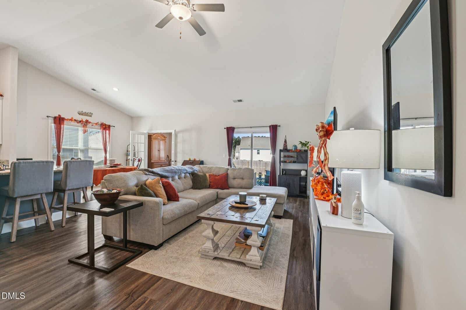 A cozy living room with a gray sectional sofa, wooden coffee table, and fall-themed decor. The space is bright with natural light from large windows and a glass door, and features an open layout with a kitchen and dining area.