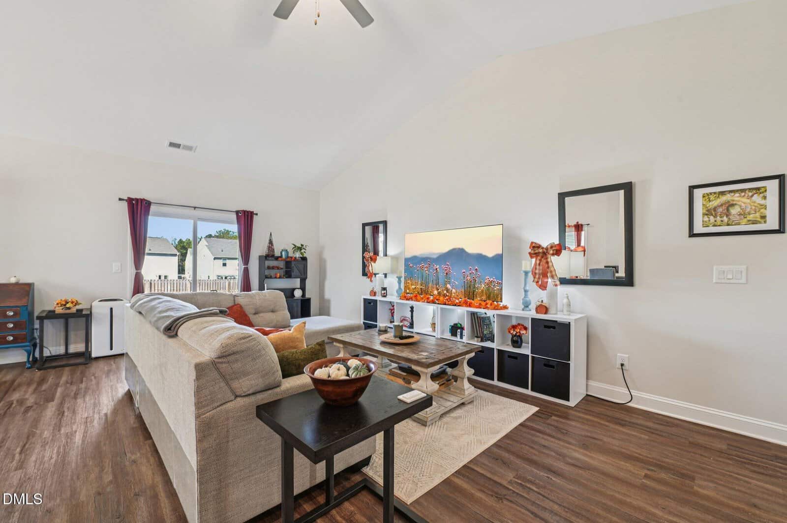 A bright, modern living room with a beige sectional sofa, a wooden coffee table, a TV on a white entertainment unit, and fall decorations. Large windows with purple curtains let in natural light. Hardwood floors throughout.