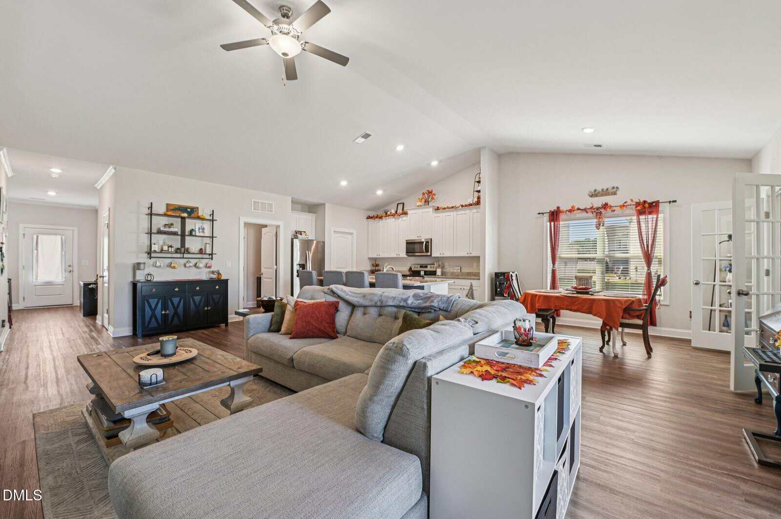 Open-concept living room and kitchen with neutral décor, a large gray sectional sofa, coffee table, dining table by the window, and fall-themed decorations throughout the space.