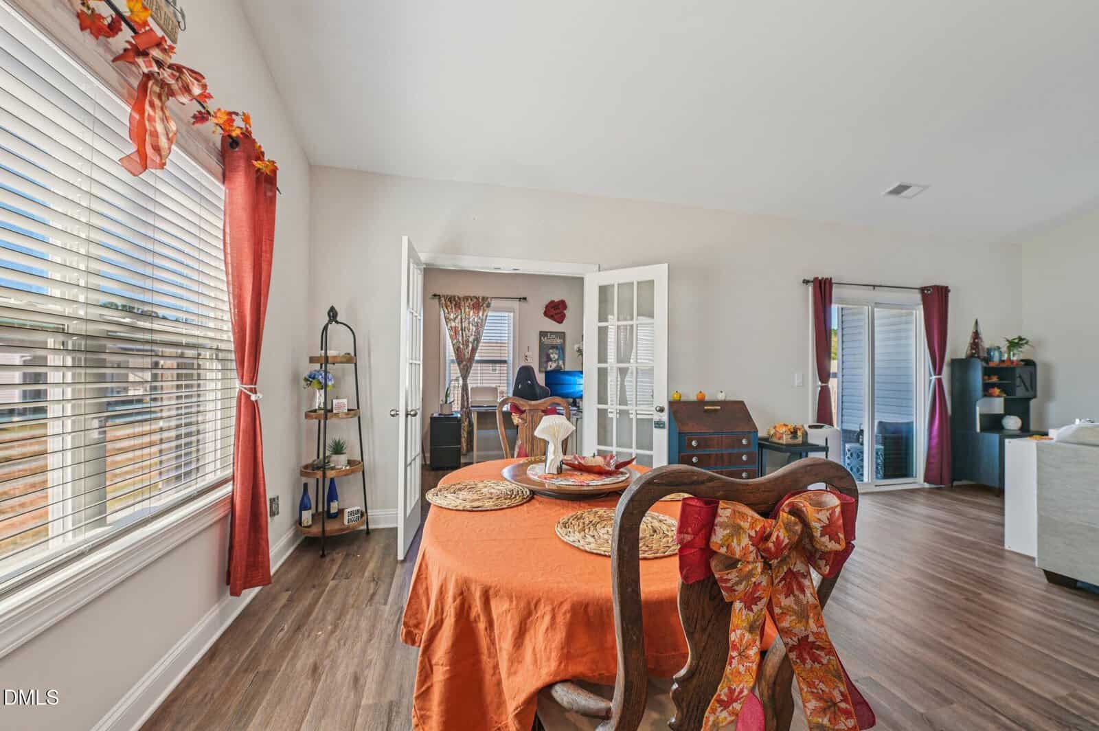 Dining room with an orange tablecloth, wooden chairs with bow decorations, and autumn-themed decor. French doors open to a study area, and large windows let in natural light. Hardwood floors run throughout the space.