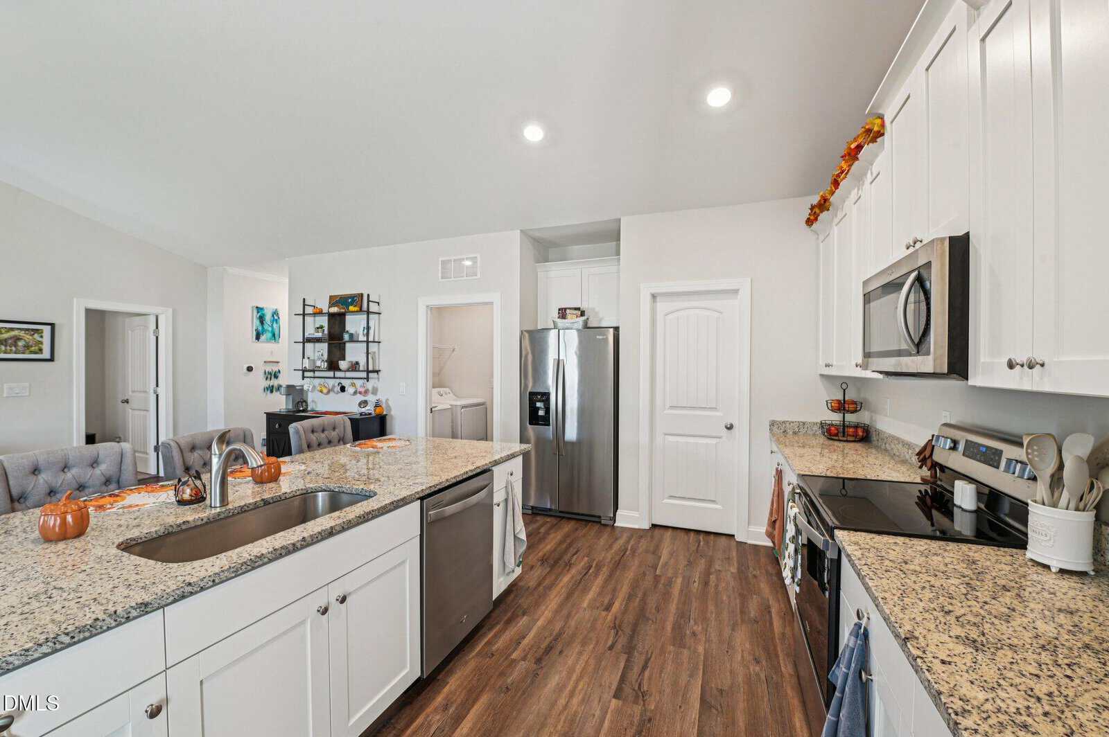 Modern kitchen with white cabinets, granite countertops, stainless steel appliances, and wood floors. Fall decorations, including small pumpkins, are on the island and above the cabinets. Dining area is visible at left.