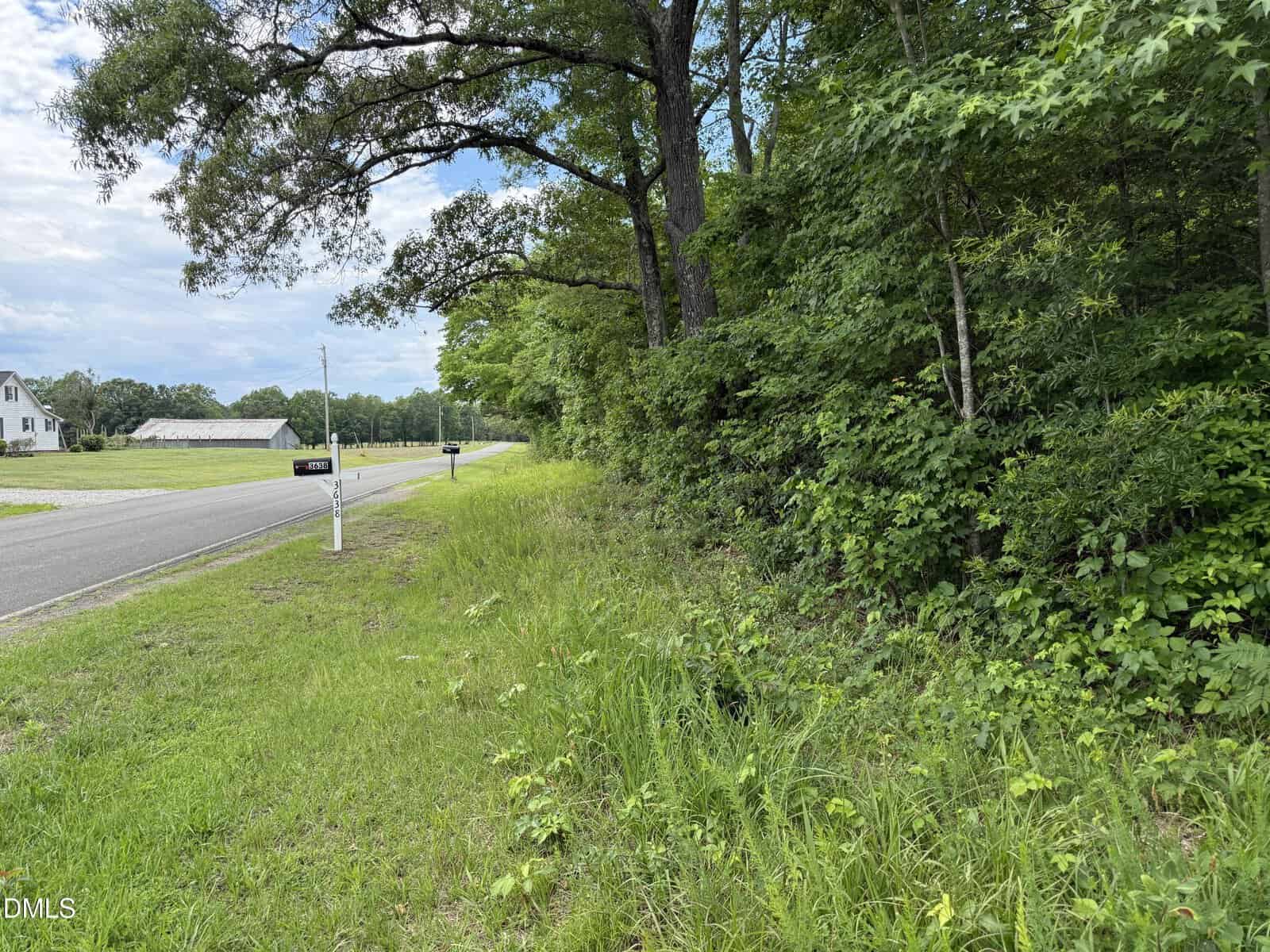 A paved road runs beside a grassy area and dense green trees. Mailboxes line the roadside, with a house and a barn visible in the distance under a partly cloudy sky.