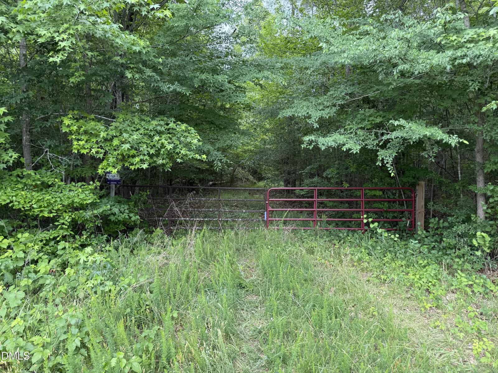 A red metal gate blocks a grassy path leading into dense green woods. There is a second silver wire gate behind it, and trees surround the area.