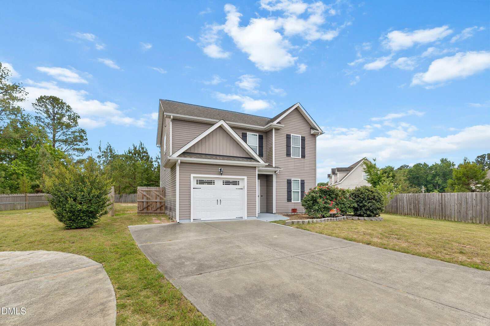 Two-story beige house with white trim and a single-car garage, set on a large lot with a concrete driveway, surrounded by grass, shrubs, and a wooden fence under a blue sky with scattered clouds.