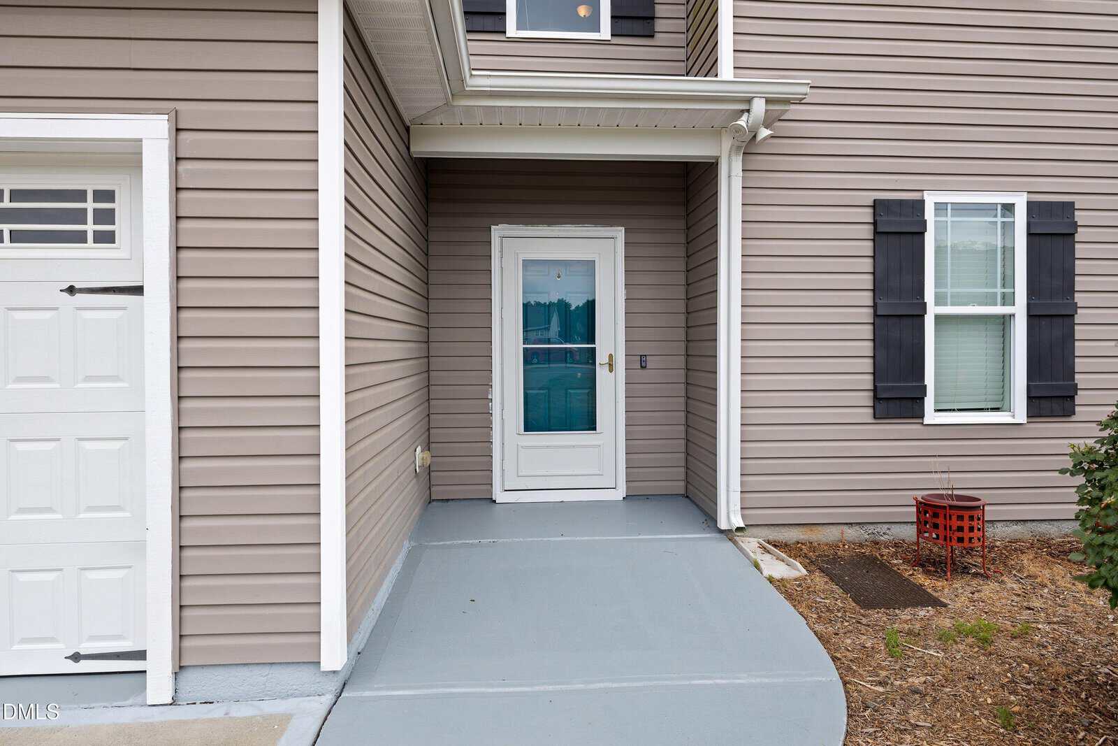 Front entryway of a house with beige siding, a white storm door, and black shuttered windows. A small porch leads to the door, and there is a red metal decorative item near a shrub on the right.