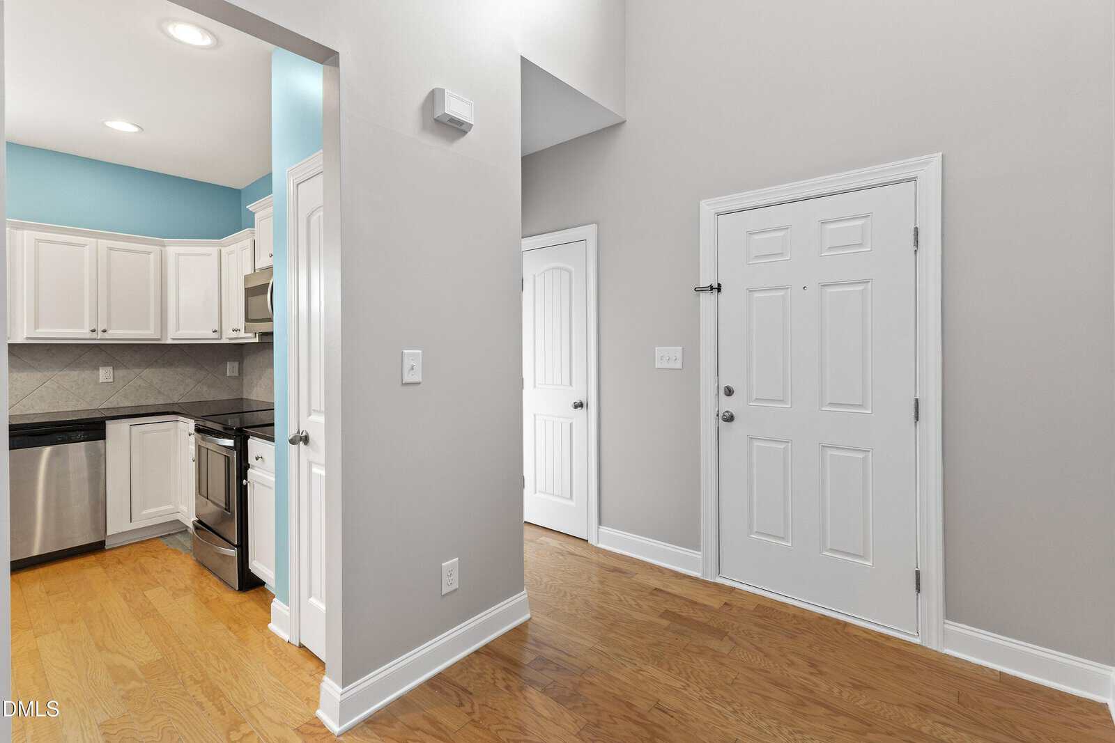 A bright entryway with light gray walls, wooden floors, and a white front door. To the left, a modern kitchen with white cabinets, stainless steel appliances, and a light blue accent wall is partially visible.