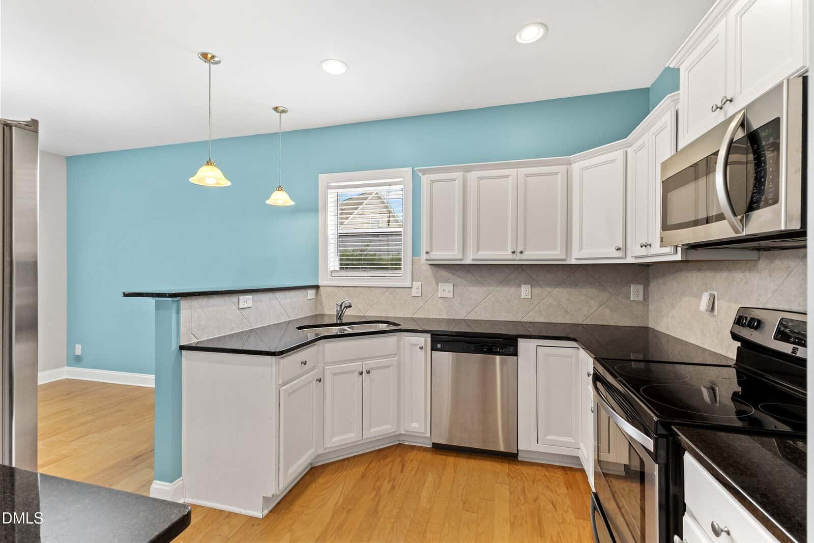 Modern kitchen with white cabinets, stainless steel appliances, black countertops, light wood flooring, blue accent wall, two pendant lights, and a window above the sink.