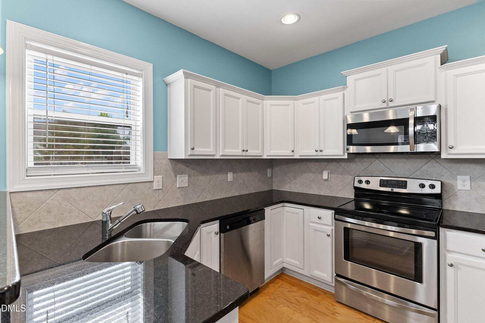 Modern kitchen with white cabinets, stainless steel appliances, black countertops, a double sink, wood flooring, and a window with blinds. The walls are painted light blue and there is geometric tile backsplash.