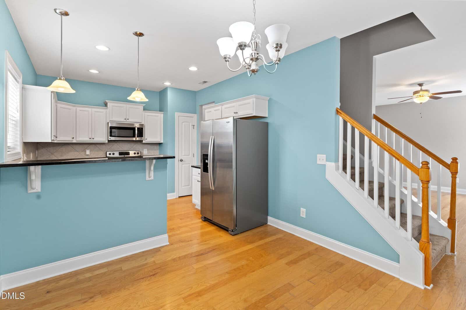 Modern kitchen with light blue walls, white cabinets, stainless steel appliances, wood floors, and an adjacent staircase with white railings and wooden handrails. A chandelier and ceiling fan are visible.