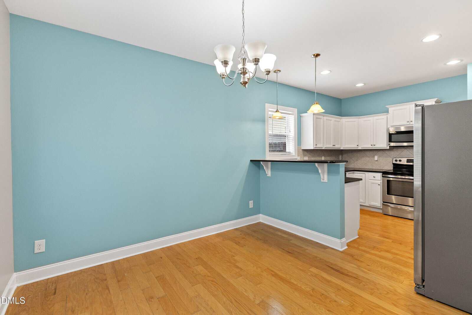 A dining area with light blue walls and wood floor, next to a kitchen with white cabinets, stainless steel appliances, granite counters, and two pendant lights. A small breakfast bar separates the spaces.