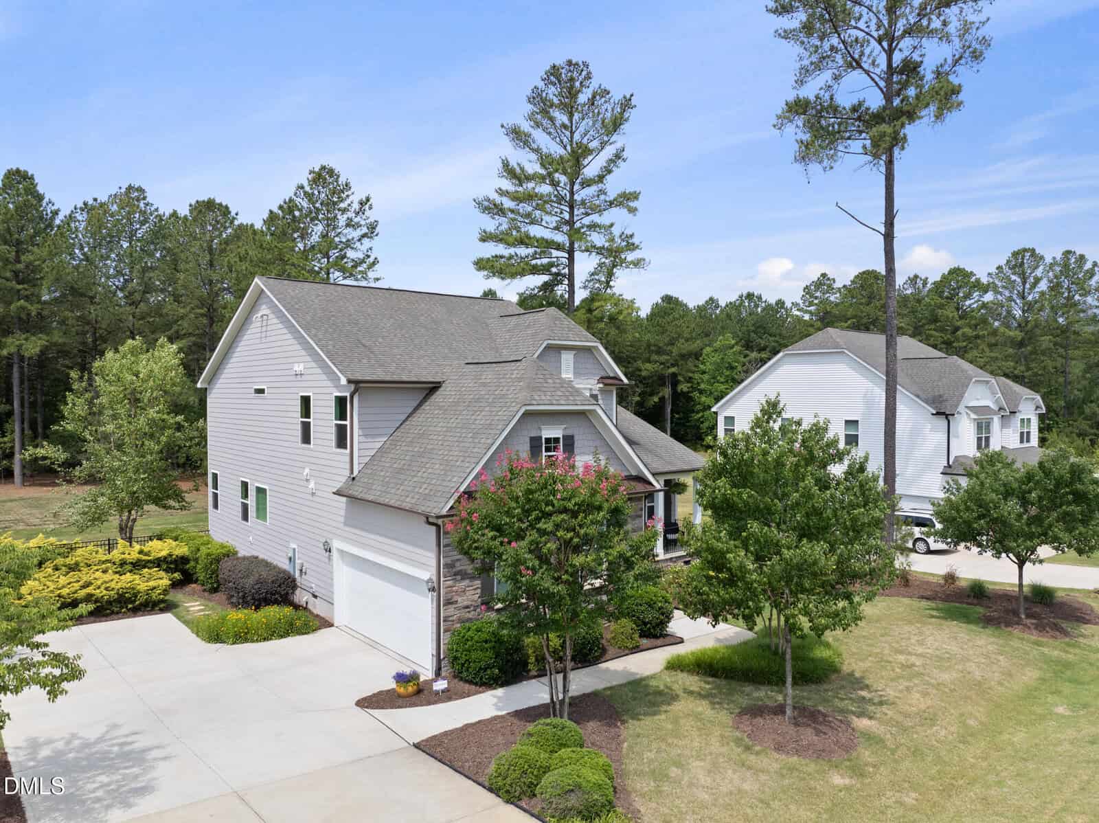 A two-story gray house with a gabled roof, white trim, and a double garage sits among tall pine trees and landscaped greenery on a spacious lot in a suburban neighborhood under a clear sky.