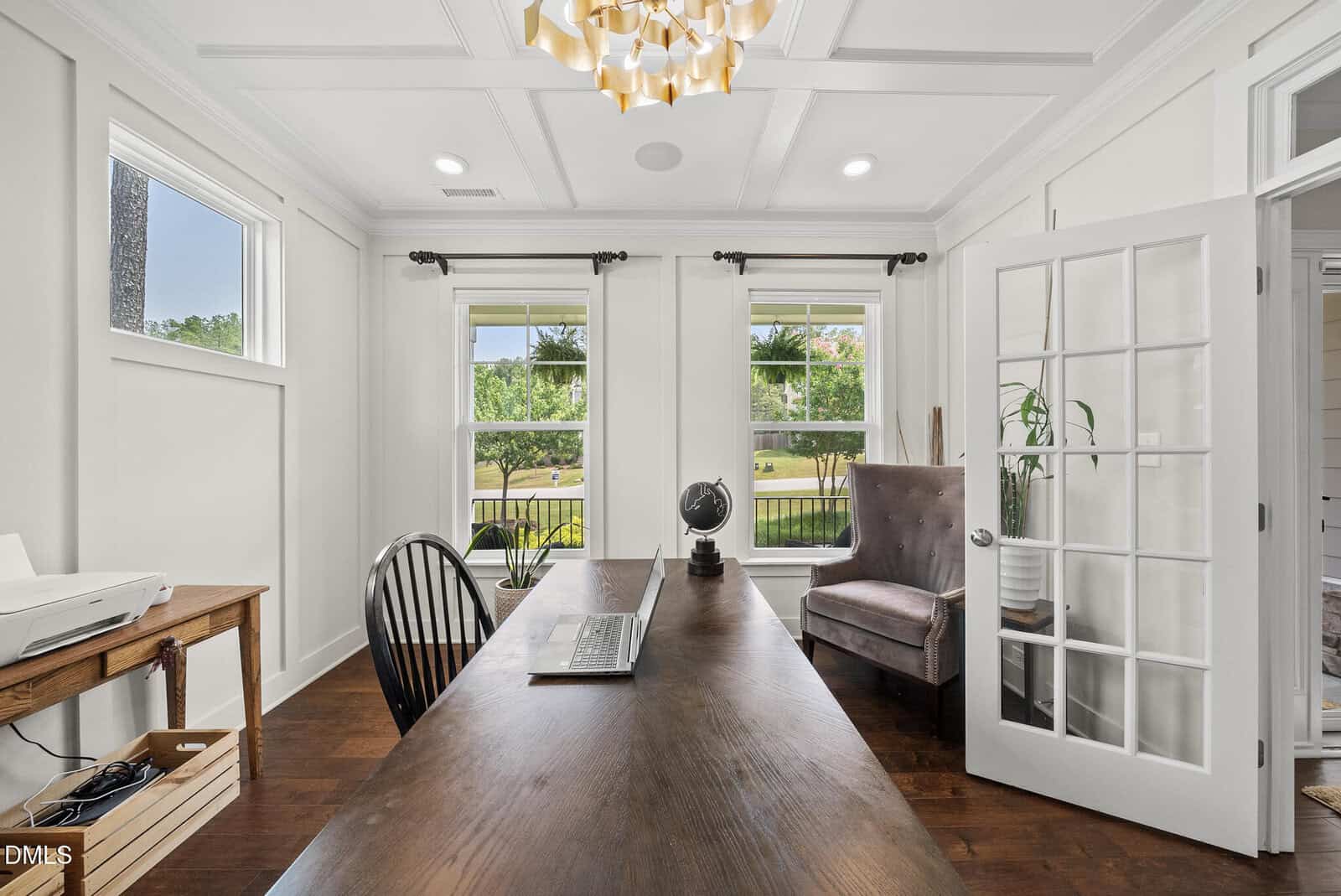 A bright home office with a wooden desk, black chair, globe, brown armchair, windows with a garden view, and French doors. A printer sits on a side table. The room has white walls and a modern chandelier.