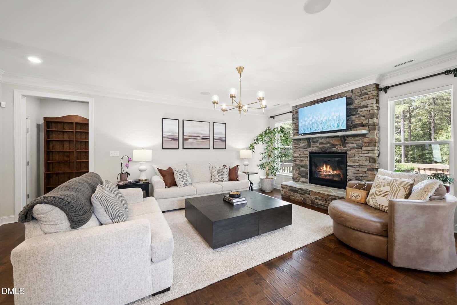 Modern living room with neutral-toned sofas, a dark coffee table, and a stone fireplace with a TV above. Large windows let in natural light, and there are plants, wall art, and bookshelves in the cozy, inviting space.