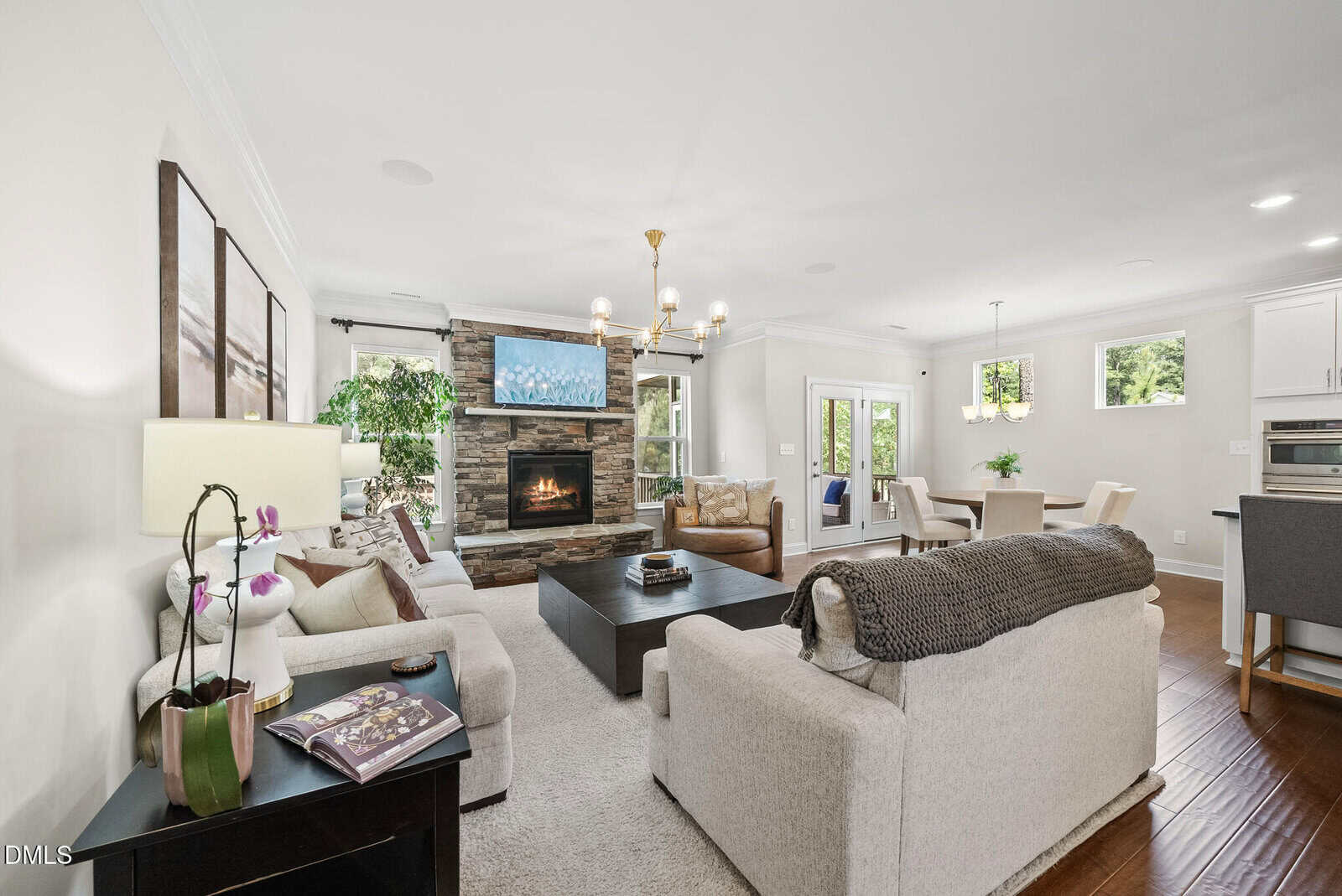 A bright, modern living room with beige sofas, a stone fireplace, large windows, a chandelier, and hardwood floors. The room opens to a dining area and kitchen, with natural light filling the space.